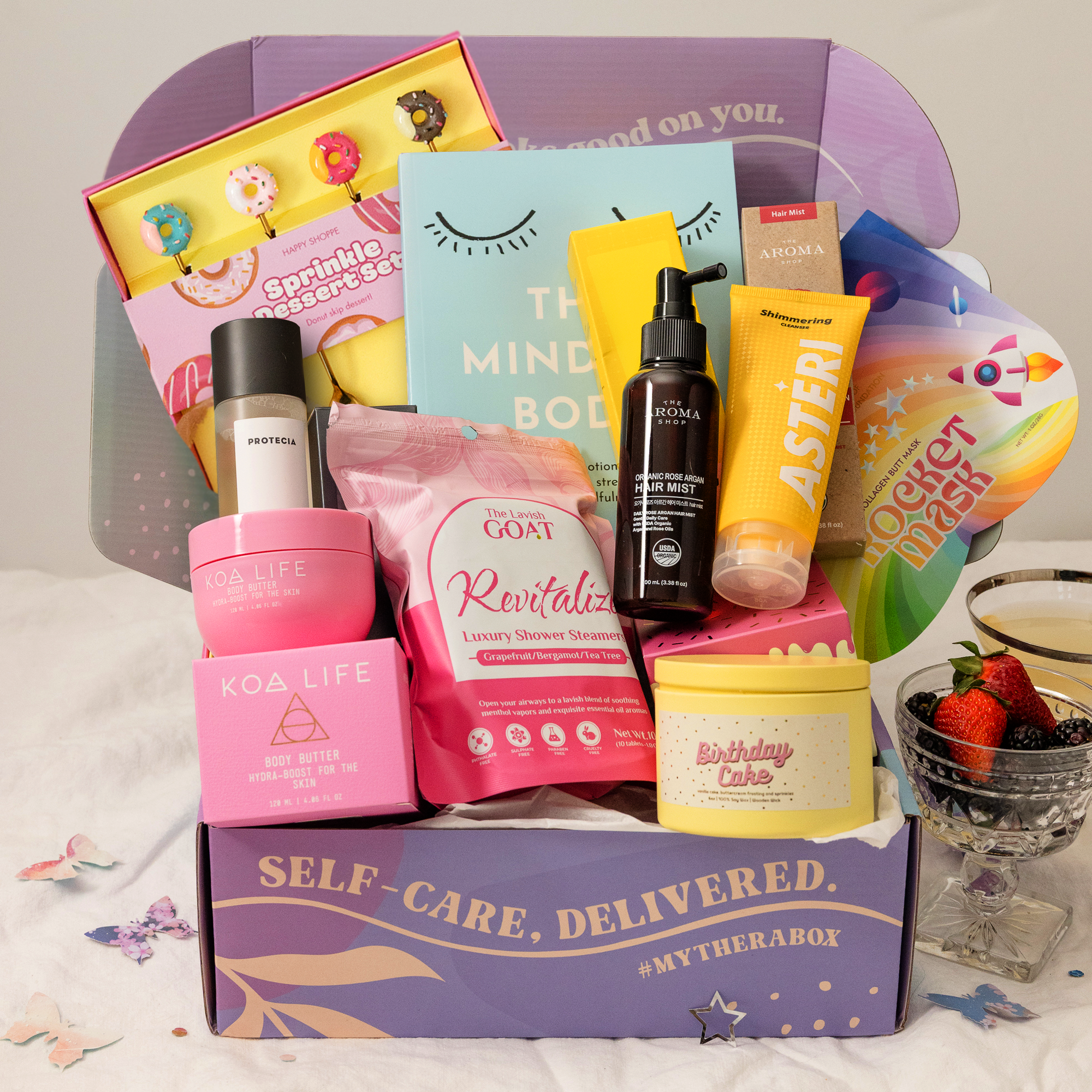 The TheraBox Cloud 9 box features KOA LIFE Hydra Boost Body Butter, BAWDY Rocket Collagen Butt Mask, PROTECIA Clarifying Toner, AYNIL Birthday Cake Candle, ALUMNI SKINCARE Shimmering Cleanser, LAVISH GOAT Revitalize Shower Steamers, THE HAPPY SHOPPE Sprinkle Dessert Set, THE AROMA SHOP Organic Rose Argan Hair Mist, and NOA BELLING X ROCKPOOL PUBLISHING’s The Mindful Body.