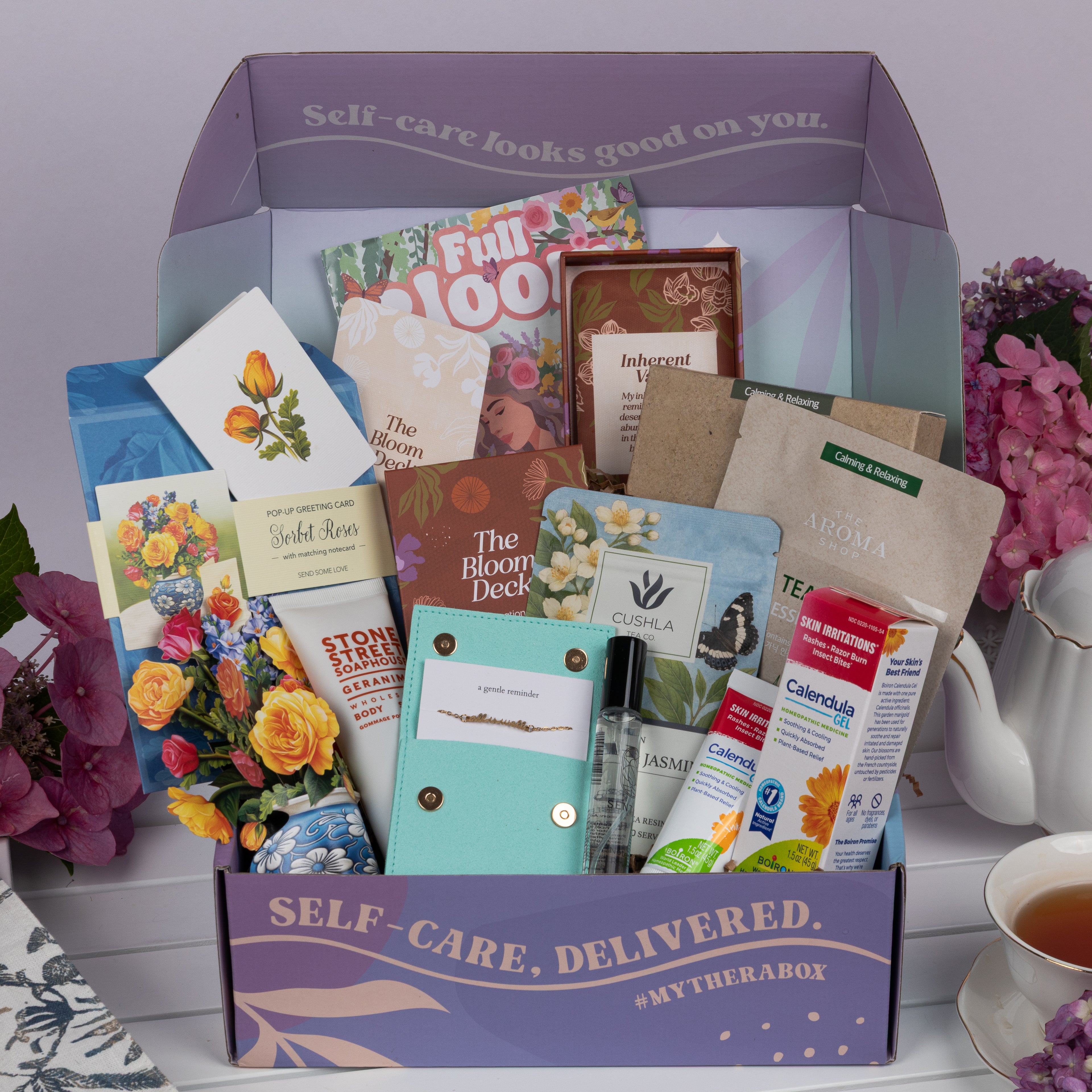 The TheraBox Full Bloom box features STONE STREET SOAPHOUSE Geranimo Body Scrub, THE AROMA SHOP Tea Tree Essence Mask 5-Pack, AYNIL CO. All Is Well Bracelet, FRESHCUT PAPER Mini Sorbet Roses, CUSHLA TEA Raw Pu-Erh & Jasmine Resin Tea, SENTIER Notes of Reverie Eau de Parfum, BOIRON USA Calendula Gel, and I KNOW COLLECTION The Bloom Deck Affirmation Cards.