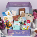 The TheraBox Full Bloom box features STONE STREET SOAPHOUSE Geranimo Body Scrub, THE AROMA SHOP Tea Tree Essence Mask 5-Pack, AYNIL CO. All Is Well Bracelet, FRESHCUT PAPER Mini Sorbet Roses, CUSHLA TEA Raw Pu-Erh & Jasmine Resin Tea, SENTIER Notes of Reverie Eau de Parfum, BOIRON USA Calendula Gel, and I KNOW COLLECTION The Bloom Deck Affirmation Cards.