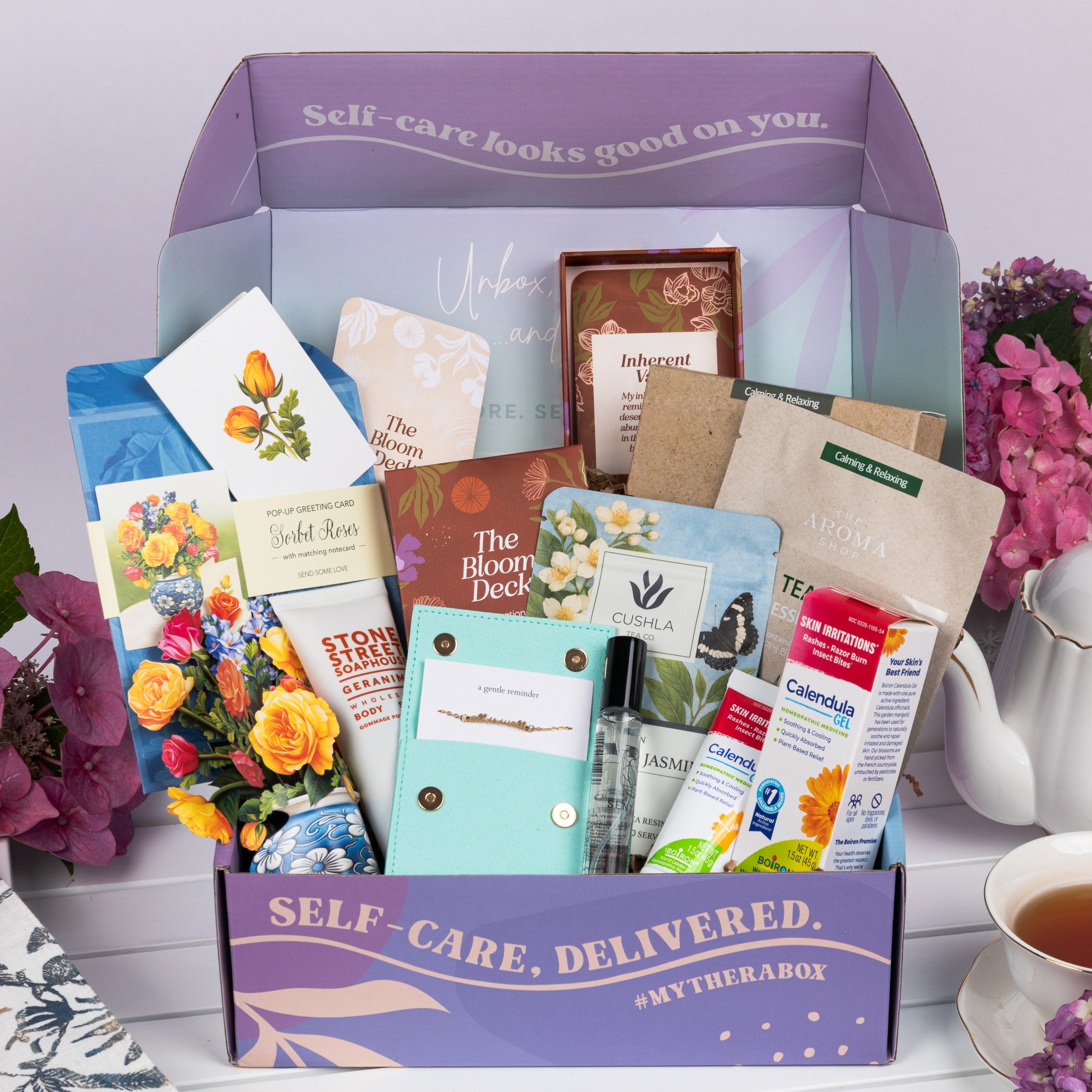 The TheraBox Full Bloom box features STONE STREET SOAPHOUSE Geranimo Body Scrub, THE AROMA SHOP Tea Tree Essence Mask 5-Pack, AYNIL CO. All Is Well Bracelet, FRESHCUT PAPER Mini Sorbet Roses, CUSHLA TEA Raw Pu-Erh & Jasmine Resin Tea, SENTIER Notes of Reverie Eau de Parfum, BOIRON USA Calendula Gel, and I KNOW COLLECTION The Bloom Deck Affirmation Cards.