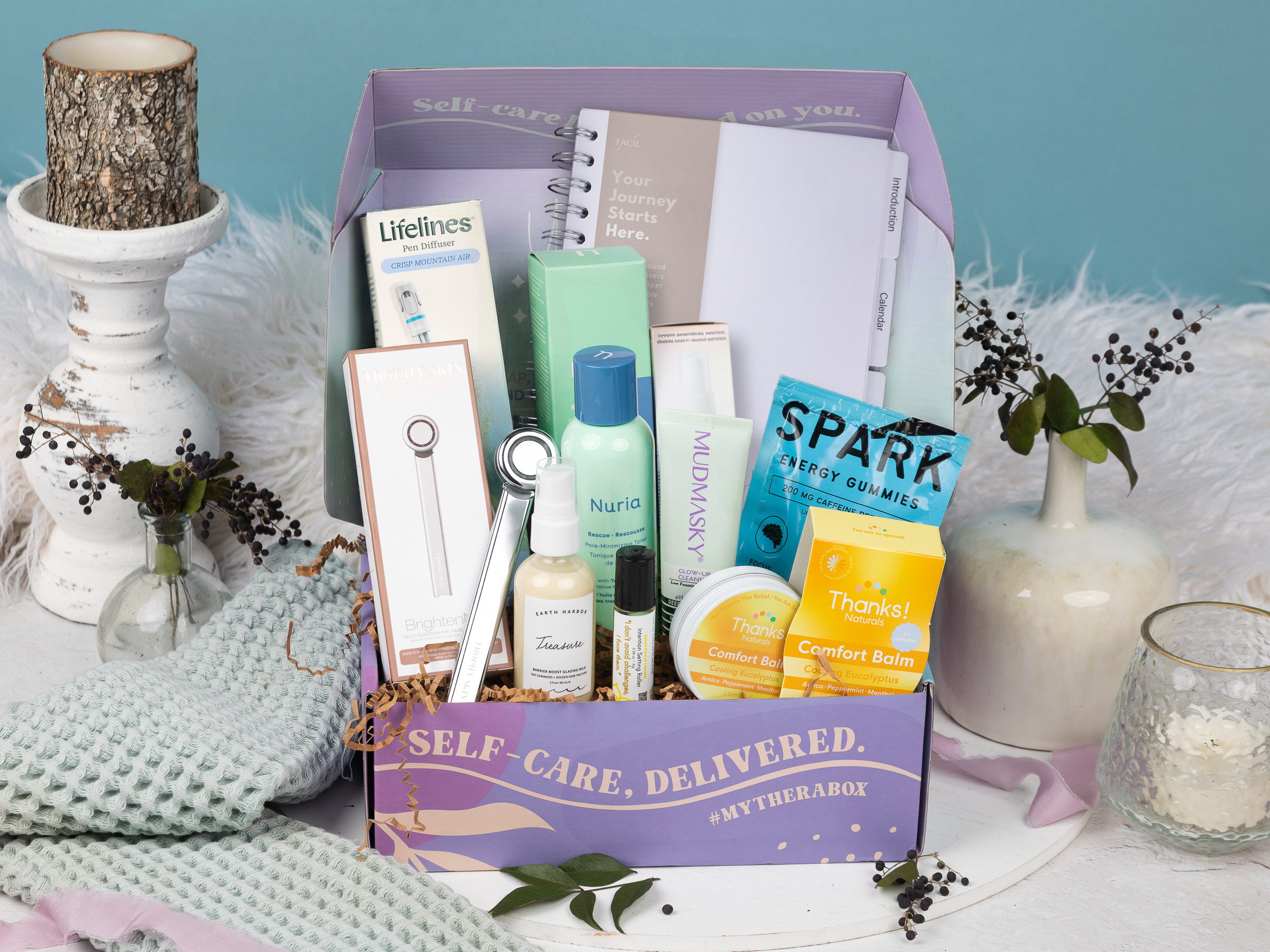 The TheraBox Glow Getter box features MUDMASKY Glow-Up Cleanser, EARTH HARBOR Treasure Barrier Boost Glazing Milk, TROPHY SKIN BrightenMD 4-In-1 Facial Device, NURIA BEAUTY Rescue Pore-Minimizing Toner, SPARK Energy Gummies, LOVE U COLLECTIVE Intention Setting Oil, THANKS! NATURALS Comfort Balm Cooling Eucalyptus, LIFELINES Crisp Mountain Air Herbal Pen Diffuser, and LA VIDA FACIL My Productivity Planner – 2026.