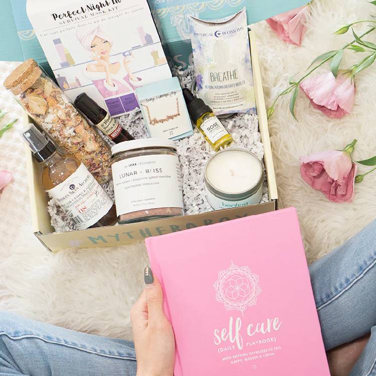 A self-care gift box assortment with a pink self-care playbook, surrounded by flowers.